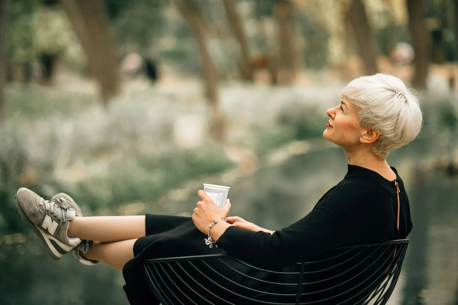 A woman with short silver hair relaxes in a garden chair with a drink, looking up at the trees.