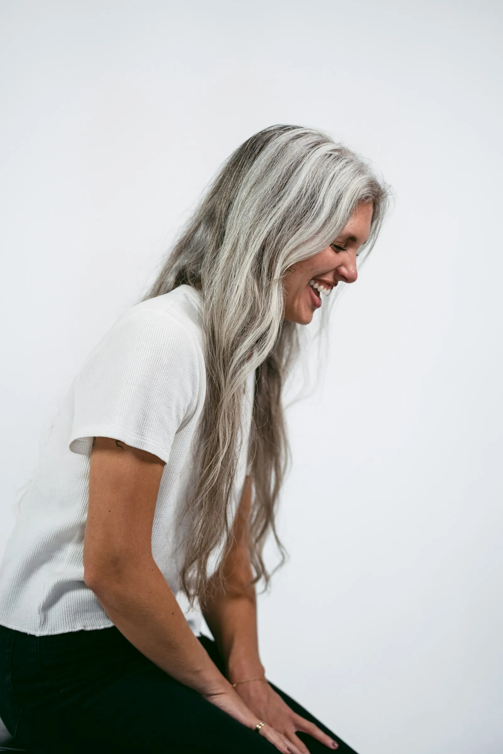 A woman with long silver hair sits in soft window light, smiling down.